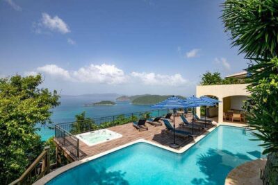 A luxury hillside villa featuring a sparkling outdoor pool and adjacent hot tub on a wood deck, with lounge chairs and blue striped umbrellas